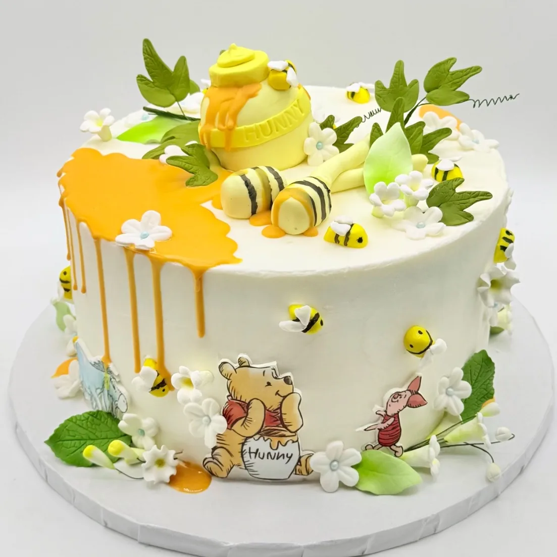 A round white buttercream cake with a golden honey drip on one side, topped with a fondant honey pot, a honey dipper, sugar bees, leaves, and white blossoms with pale blue centers, with printed storybook bear and friend characters decorating the sides.