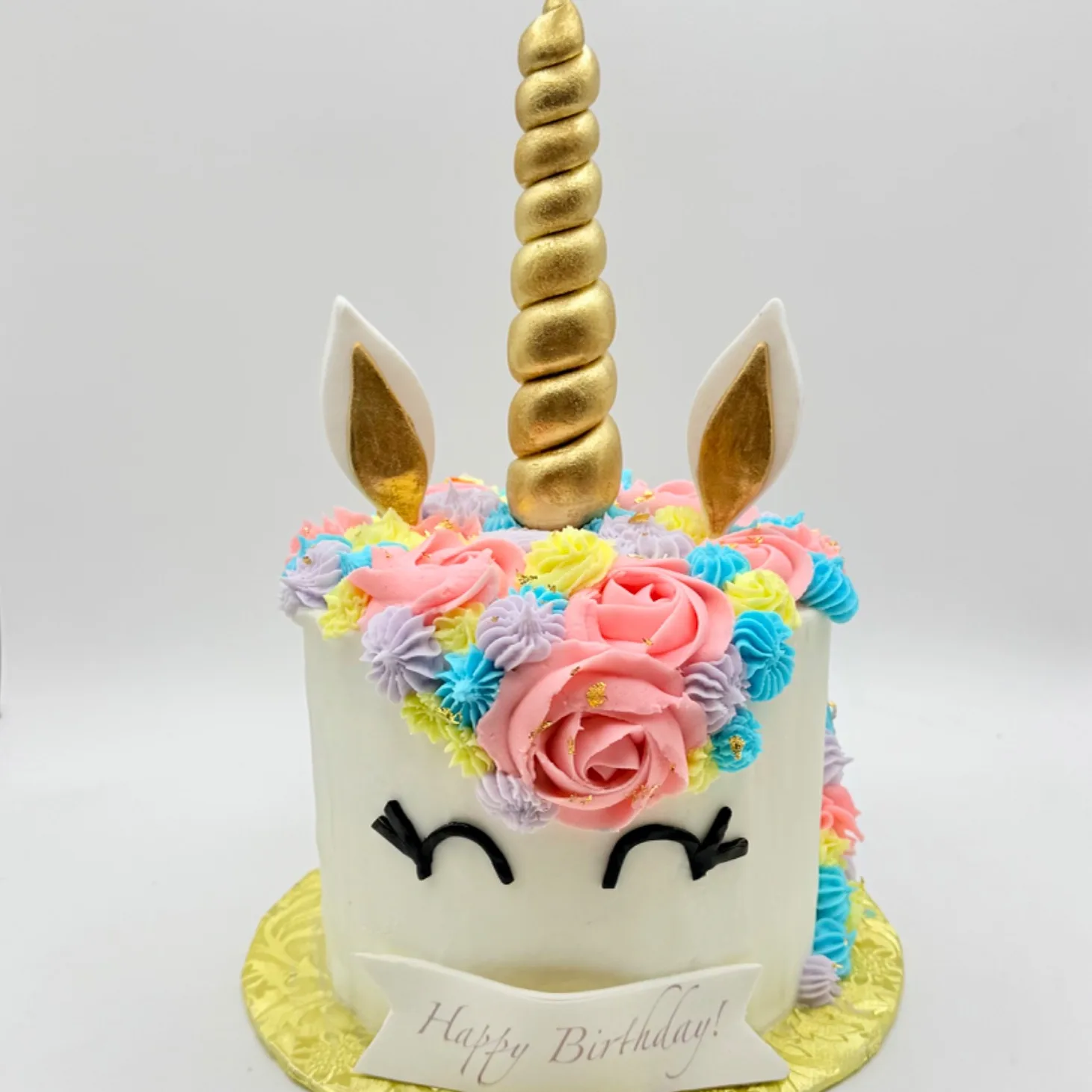 A small round unicorn cake with a gold spiral horn, gold-lined white ears, closed eyelash eyes, and a crown of pink, lavender, blue, and yellow piped flowers, with a "Happy Birthday" banner on the front, displayed on a gold cake board.