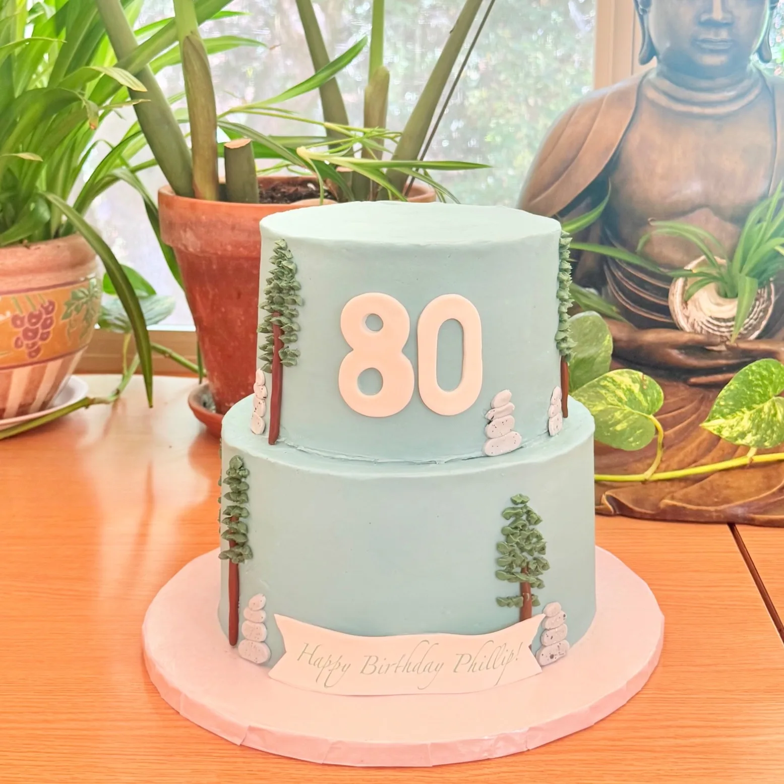 A two tier sage green buttercream cake decorated with hand piped pine trees and stacked stone cairns, a white fondant "80," and a banner reading "Happy Birthday Phillip," displayed on a table near potted plants.