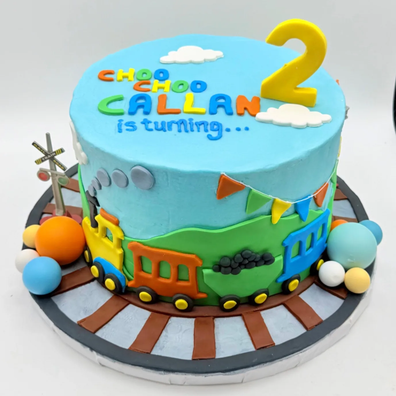 Round two tier birthday cake in sky blue buttercream with piped clouds and a hand lettered message, a yellow fondant number two, a colorful fondant train with a coal car circling the base, a small railroad crossing sign topper, and a cake board painted to look like railroad tracks.