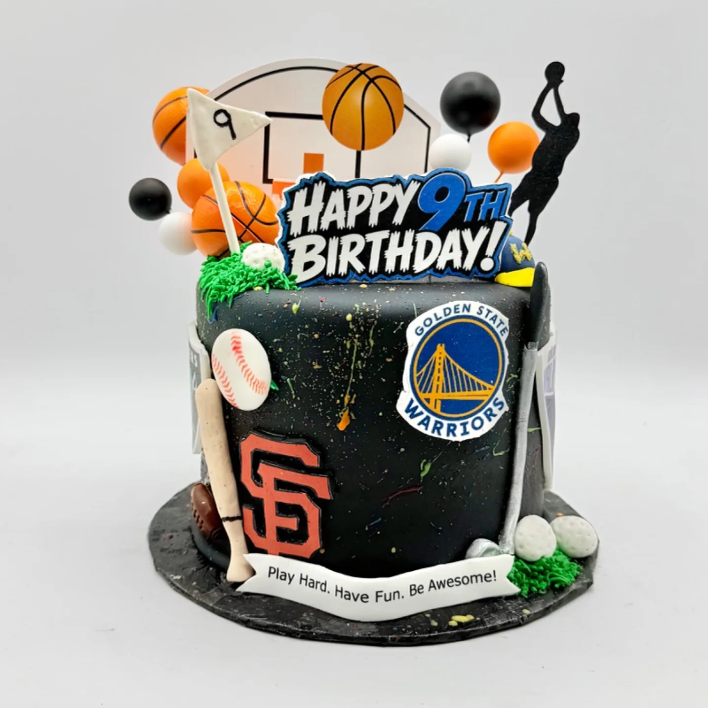 A black paint-splattered cake decorated with sports team crests, a baseball and bat, mini basketballs, golf balls, a football, balloons, and a Happy 9th Birthday topper, photographed against a plain gray background.