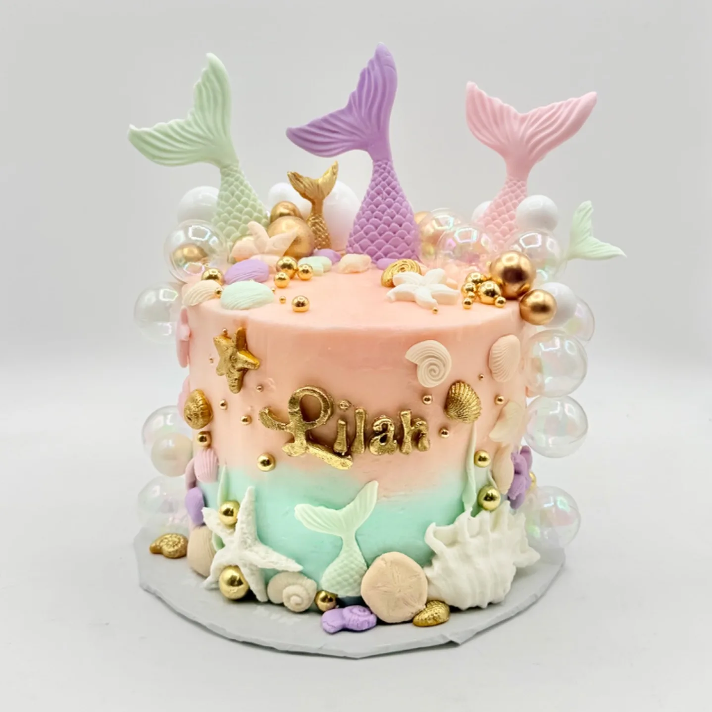 A round cake in peach and mint ombre buttercream topped with three mermaid tails in mint, lavender, and pink, surrounded by gold shells, starfish, and iridescent bubble decorations, with the name Lilah piped in gold across the front.