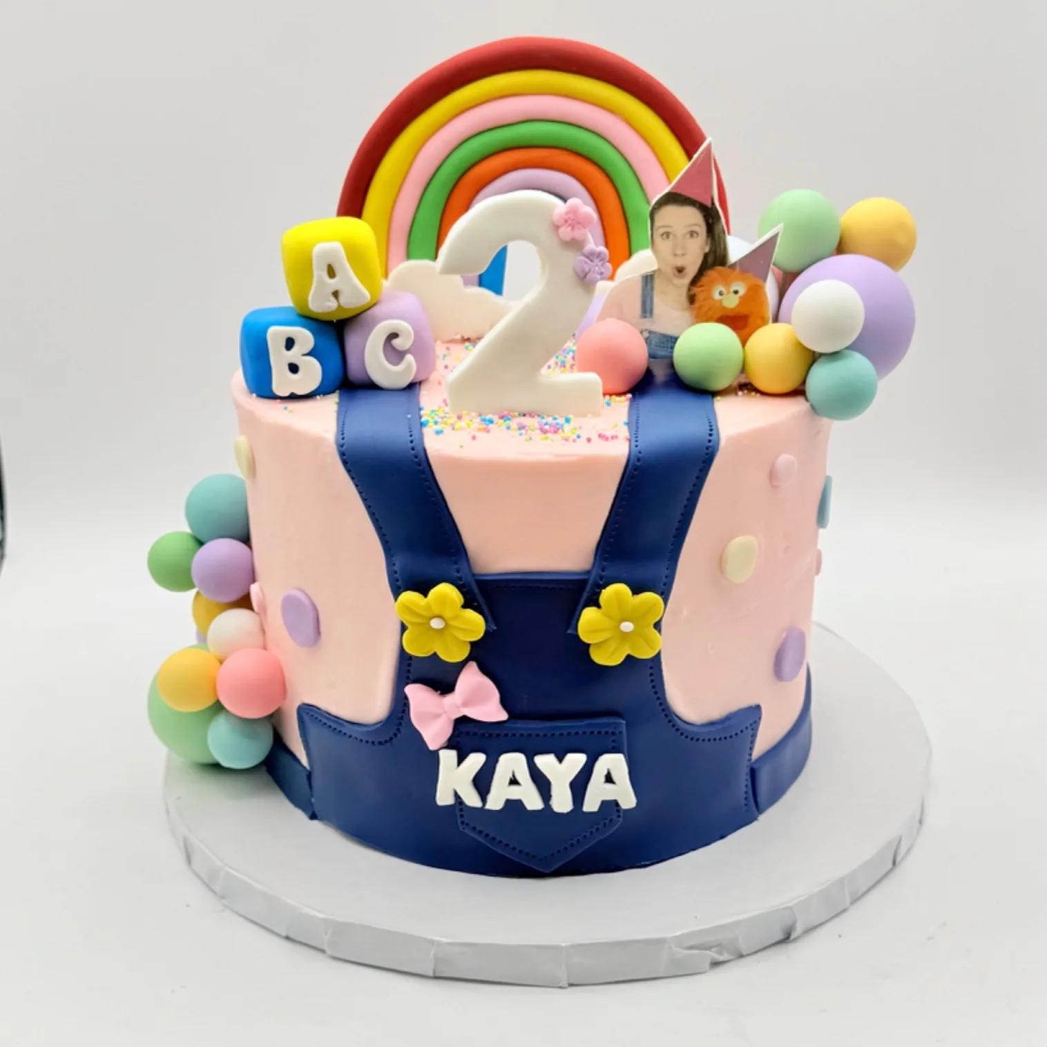 Round pink buttercream cake designed to look like a figure wearing blue denim overalls with a name plaque reading Kaya, topped with a fondant rainbow, alphabet blocks spelling ABC, a number 2, a small orange puppet character, and pastel sprinkle balls along one side.