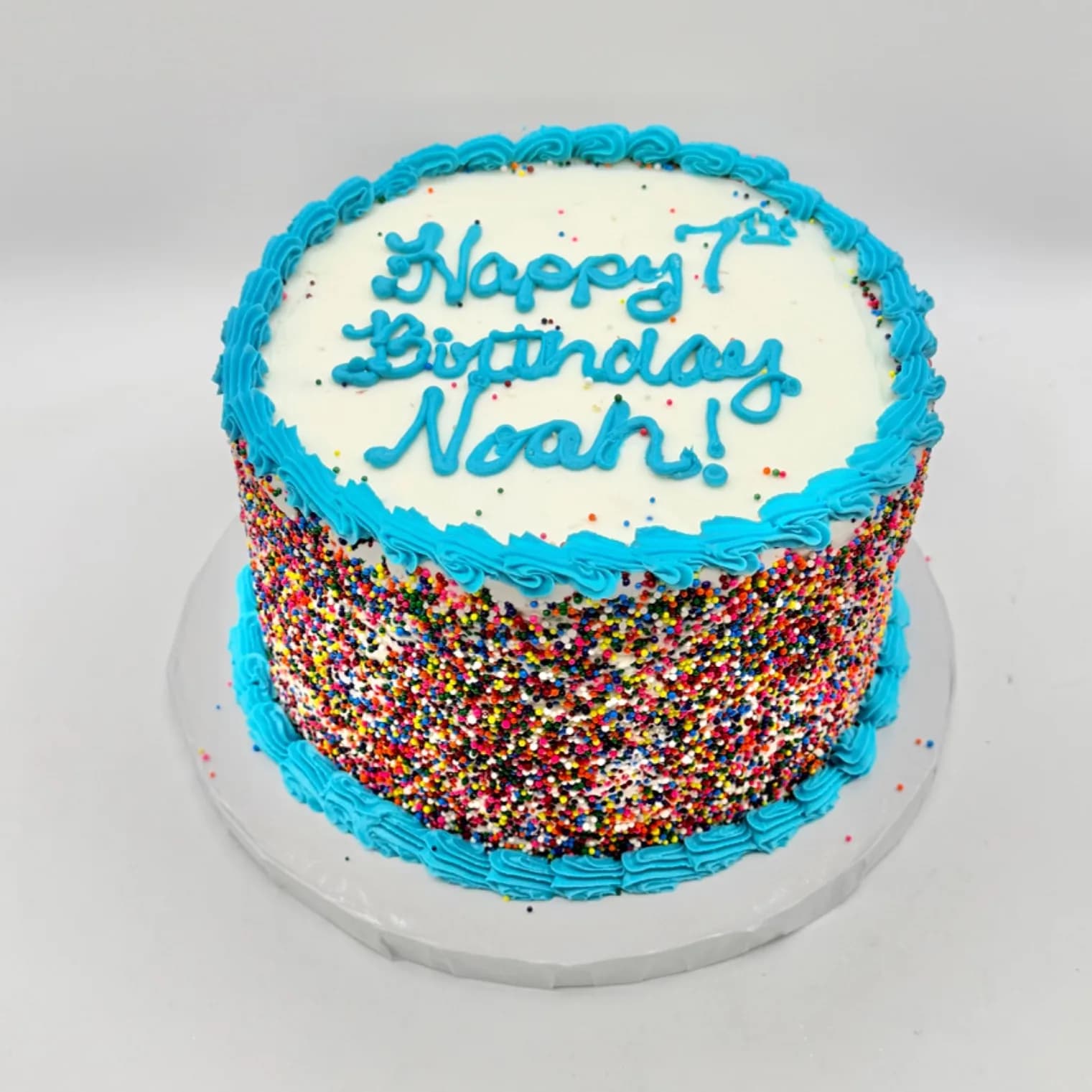 Round white buttercream cake with a rainbow sprinkle covered lower half, a teal piped border, and "Happy 7th Birthday Noah!" hand lettered in teal on the plain white top.