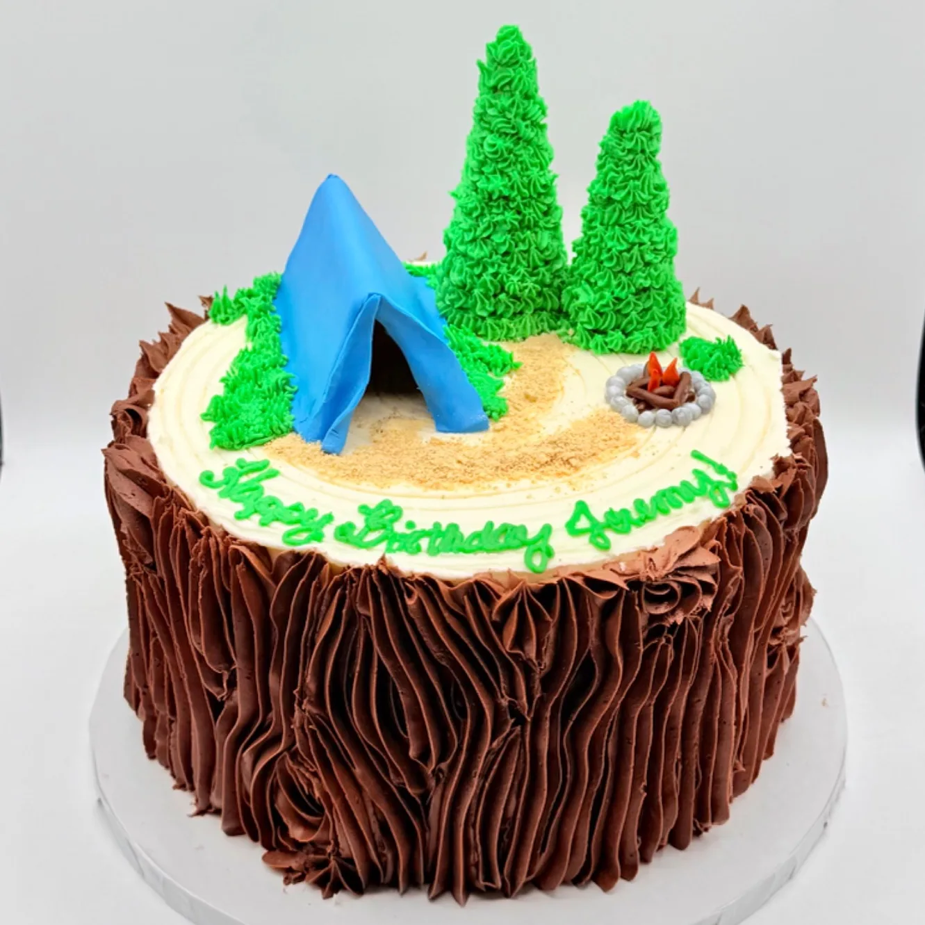 Round cake decorated to look like a chocolate buttercream tree stump, topped with a fondant camping tent, two piped pine trees, a miniature campfire, and a graham crumb path, with a hand piped birthday message on the top surface.
