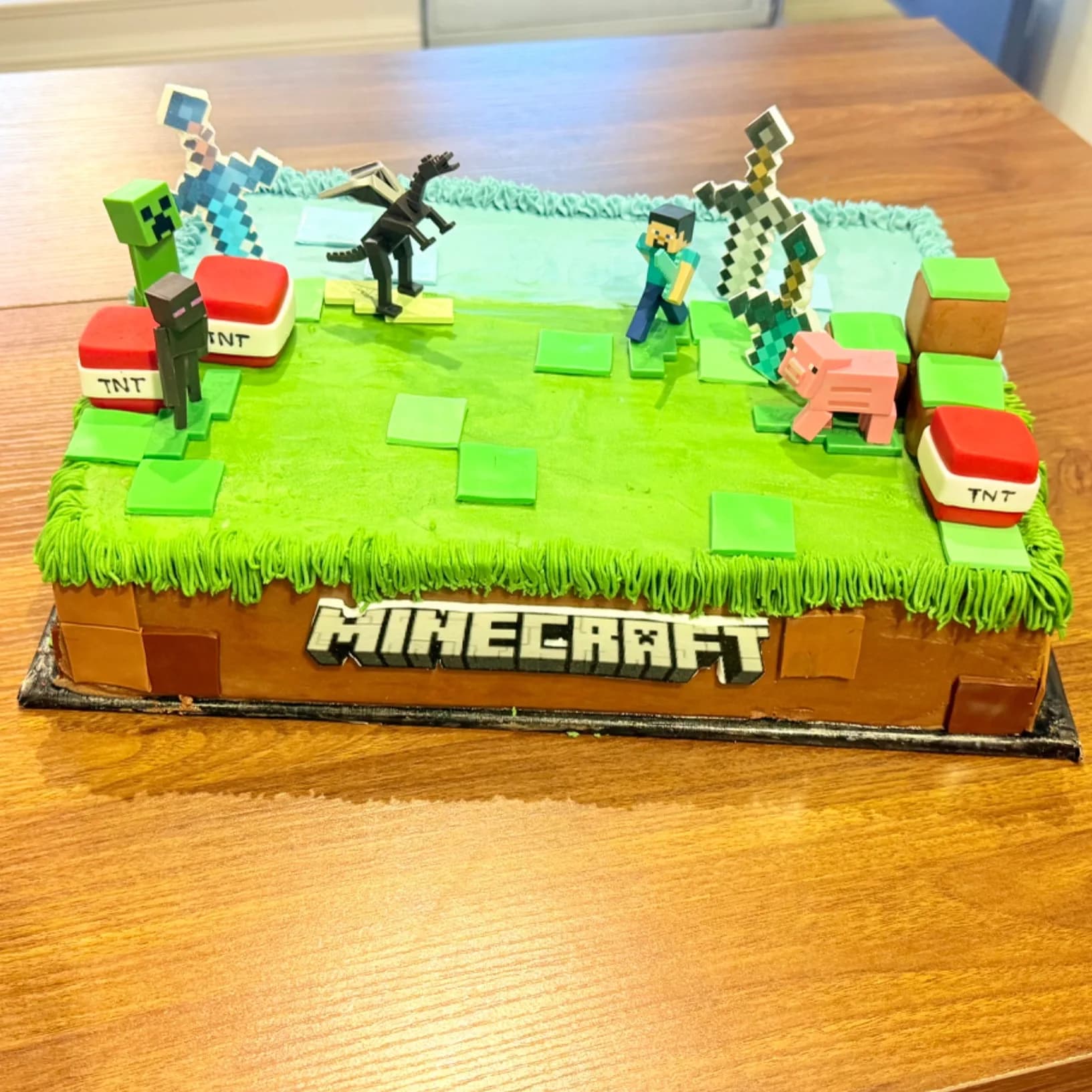 A rectangular sheet cake decorated as a blocky green and brown landscape with fondant grass squares, TNT blocks, pixelated sword cutouts, a block-letter name plaque on the front, and several small blocky character figures arranged on top.