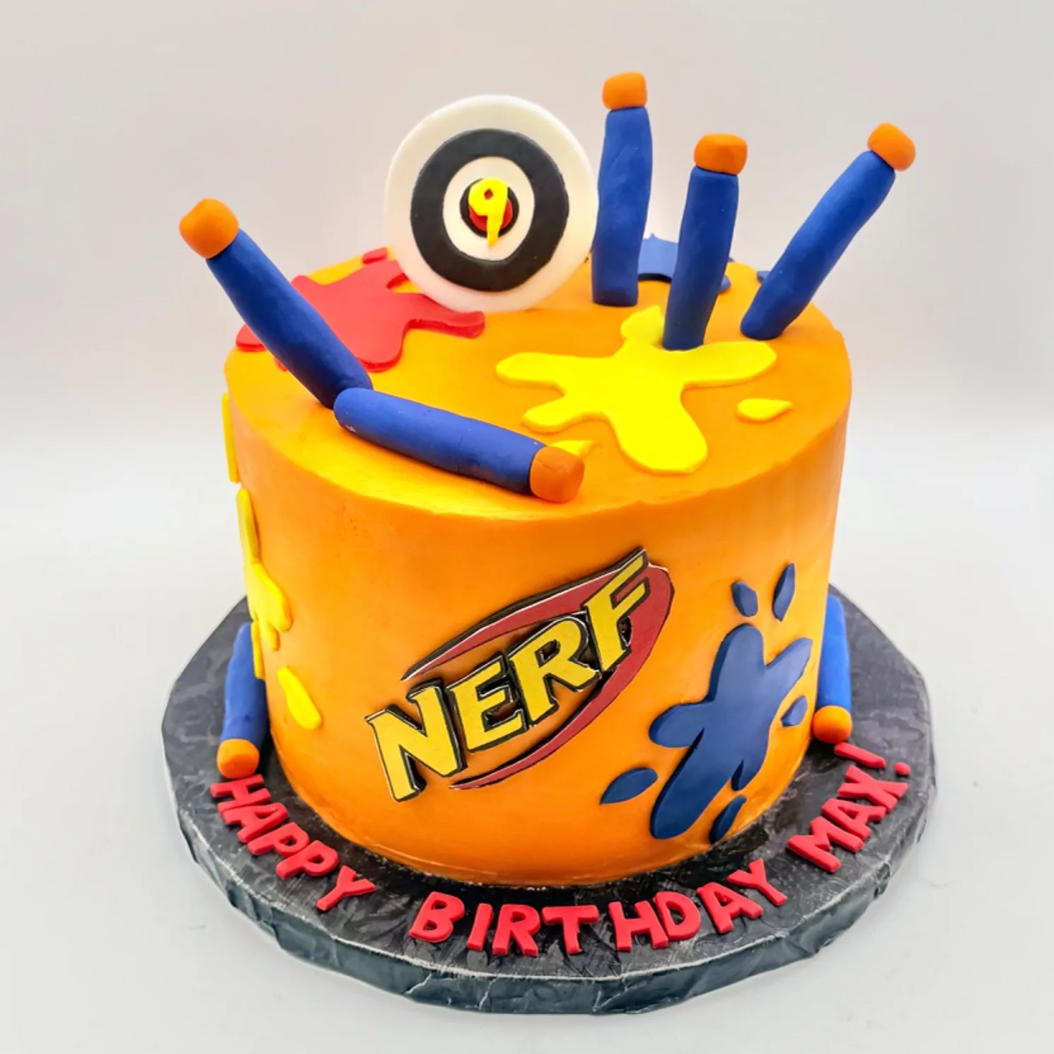 A round orange fondant birthday cake topped with blue foam dart blasters and a round black and white target with a yellow number nine, decorated with red, yellow and blue paint splatter shapes and a dart logo on the side, with "Happy Birthday Max!" piped in red on the dark cake board.