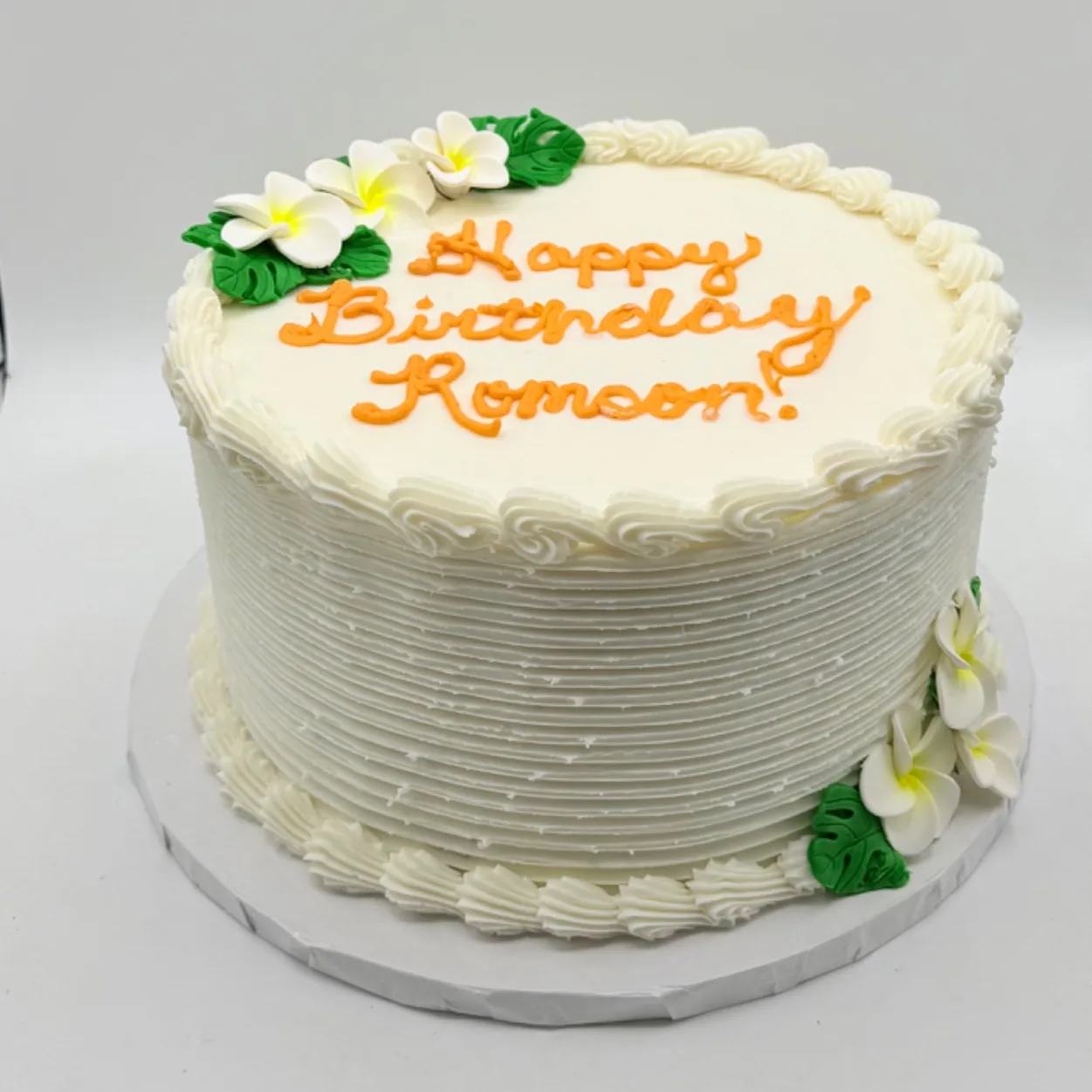 Round white buttercream cake with a textured comb finish on the sides, sugar plumeria flowers and monstera leaves in two corners, and "Happy Birthday Romson!" piped across the top in orange script.