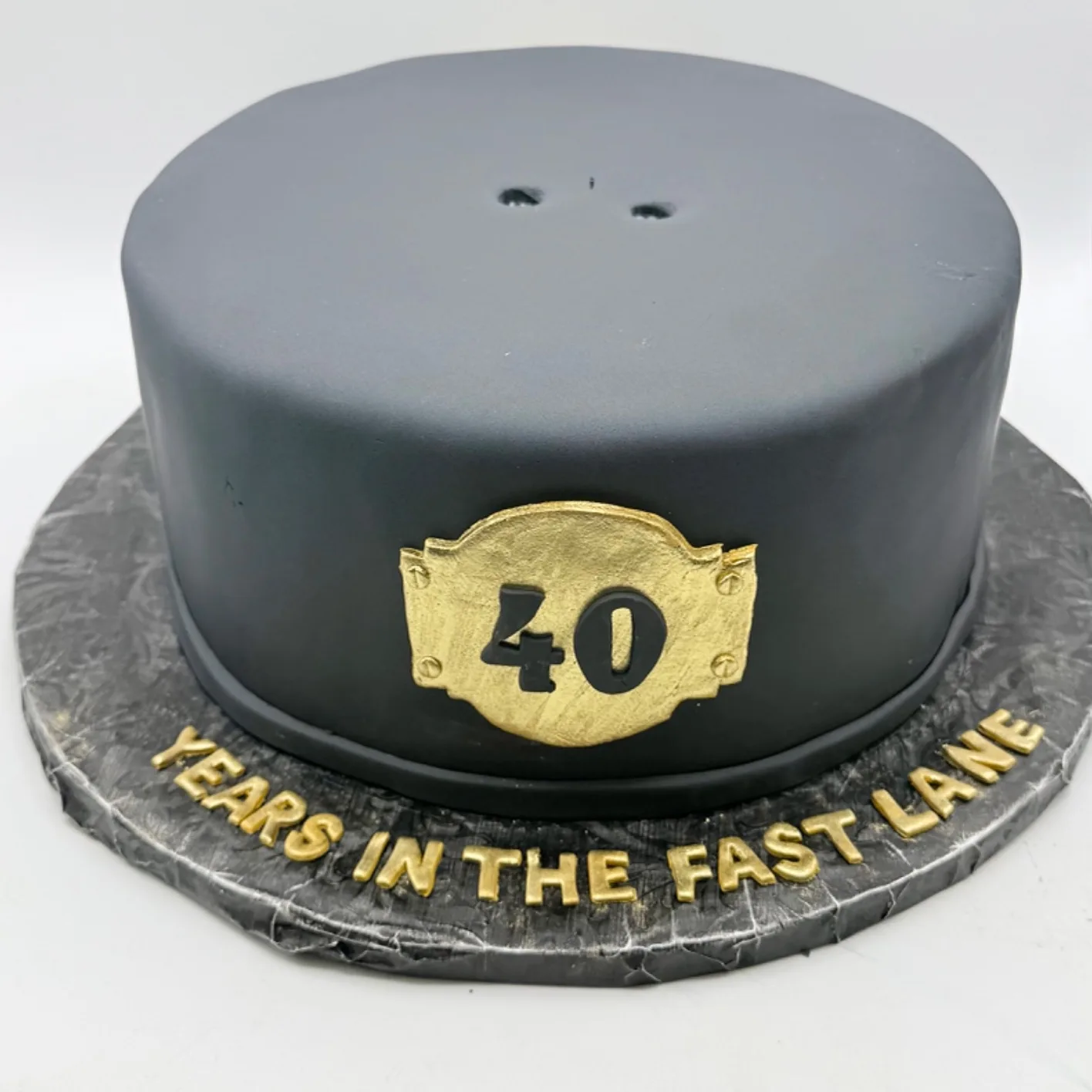 A round black fondant birthday cake with a hand painted gold plaque reading "40" with bolt details, set on a slate textured board with "Years in the Fast Lane" piped in gold.