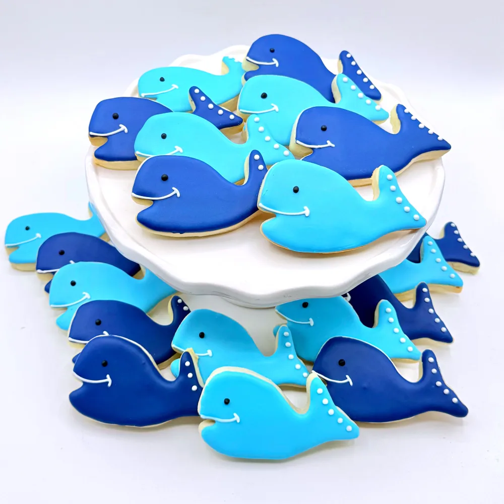 Whale cookies iced in light and dark blue with white dot details, piled on a white stand