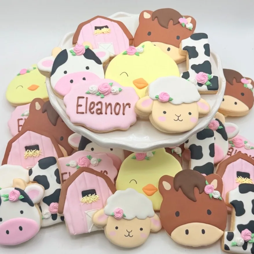 A tiered display of decorated sugar cookies shaped like farm animals, a pink barn, a cow print number one, and a name plaque reading Eleanor, all finished in pastel colors with piped rosette accents.