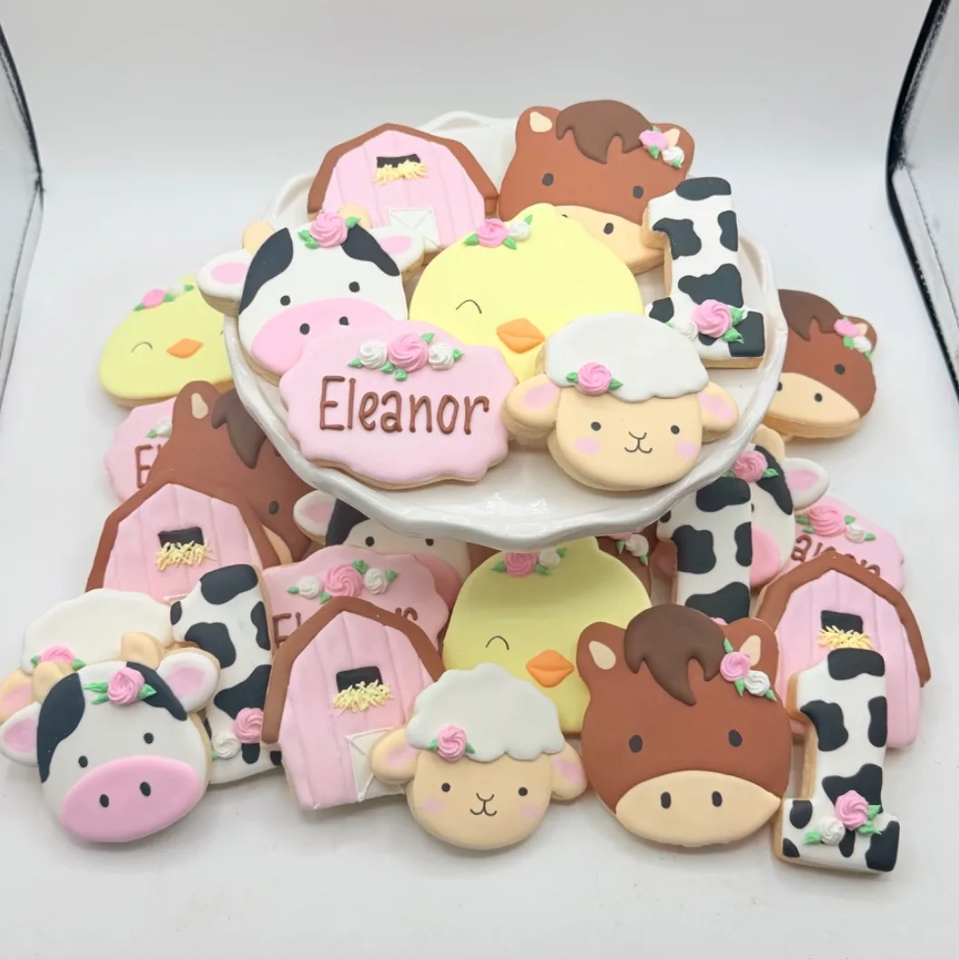 A tiered display of decorated sugar cookies shaped like farm animals, a pink barn, a cow print number one, and a name plaque reading Eleanor, all finished in pastel colors with piped rosette accents.