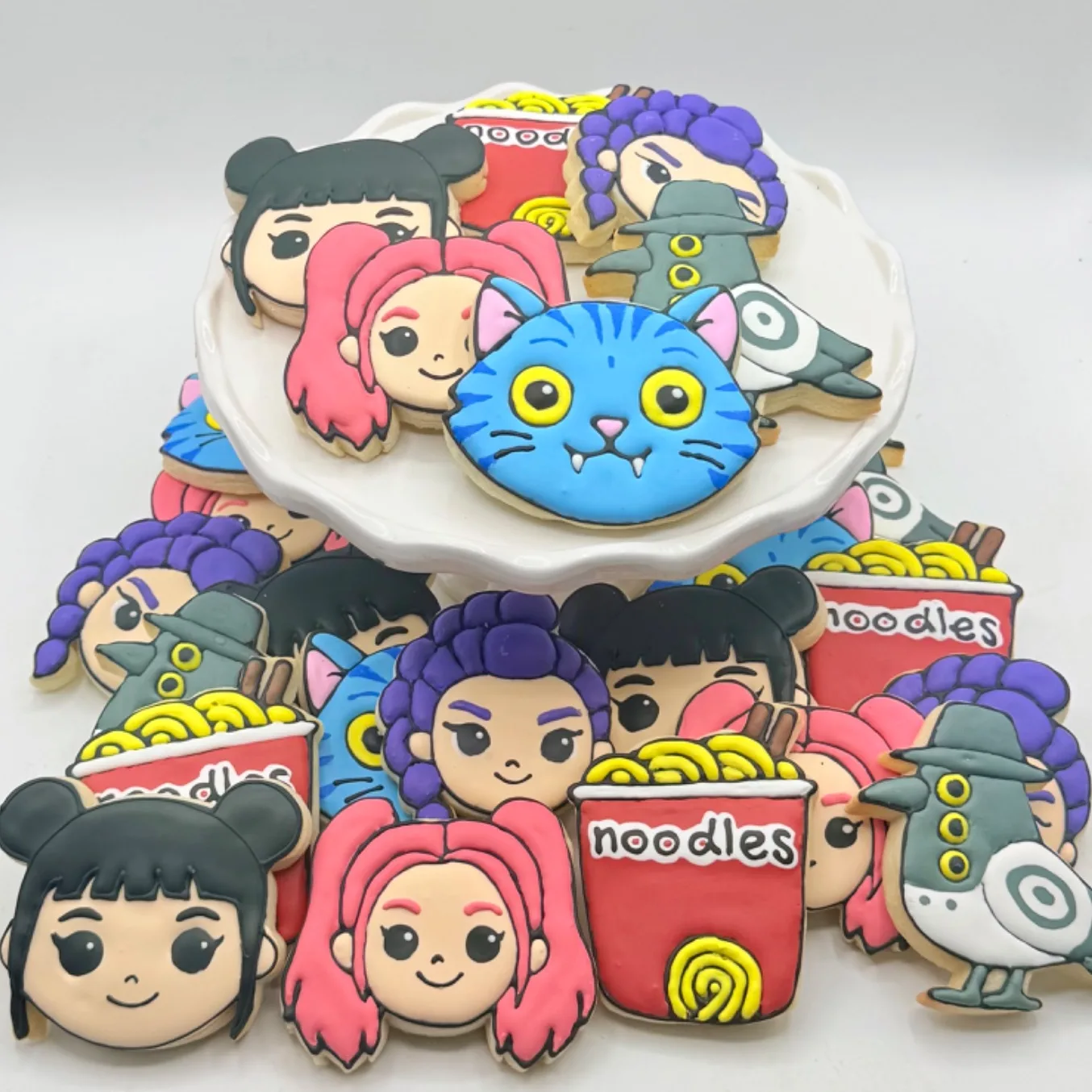 A platter of decorated sugar cookies from KPop Demon Hunters, three character faces, a blue fanged cat, a gray bird with dark rings around its eyes, and a red cup of noodles with yellow ramen and chopsticks, finished in bright hand-painted icing.
