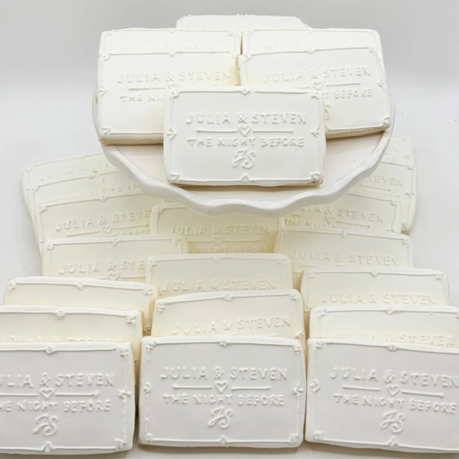 A stack of rectangular gluten free sugar cookies in white icing, each embossed with a couple's names, "the night before," and their monogram inside a decorative border, arranged on a white platter.