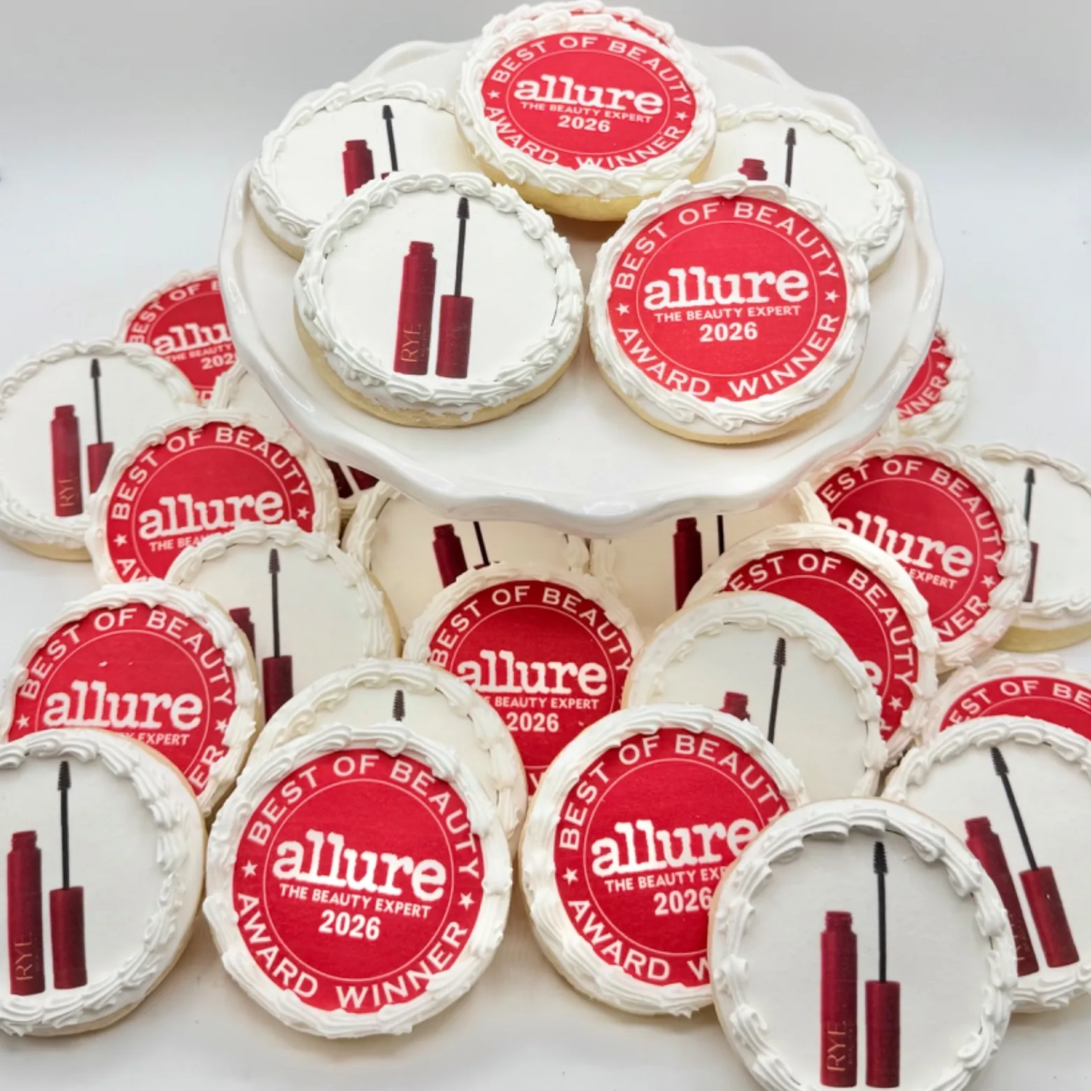 A tiered display of round white buttercream cookies, some printed with a red "Best of Beauty" award seal and others hand piped with an illustration of a mascara tube and wand, arranged on a white cake stand and table.