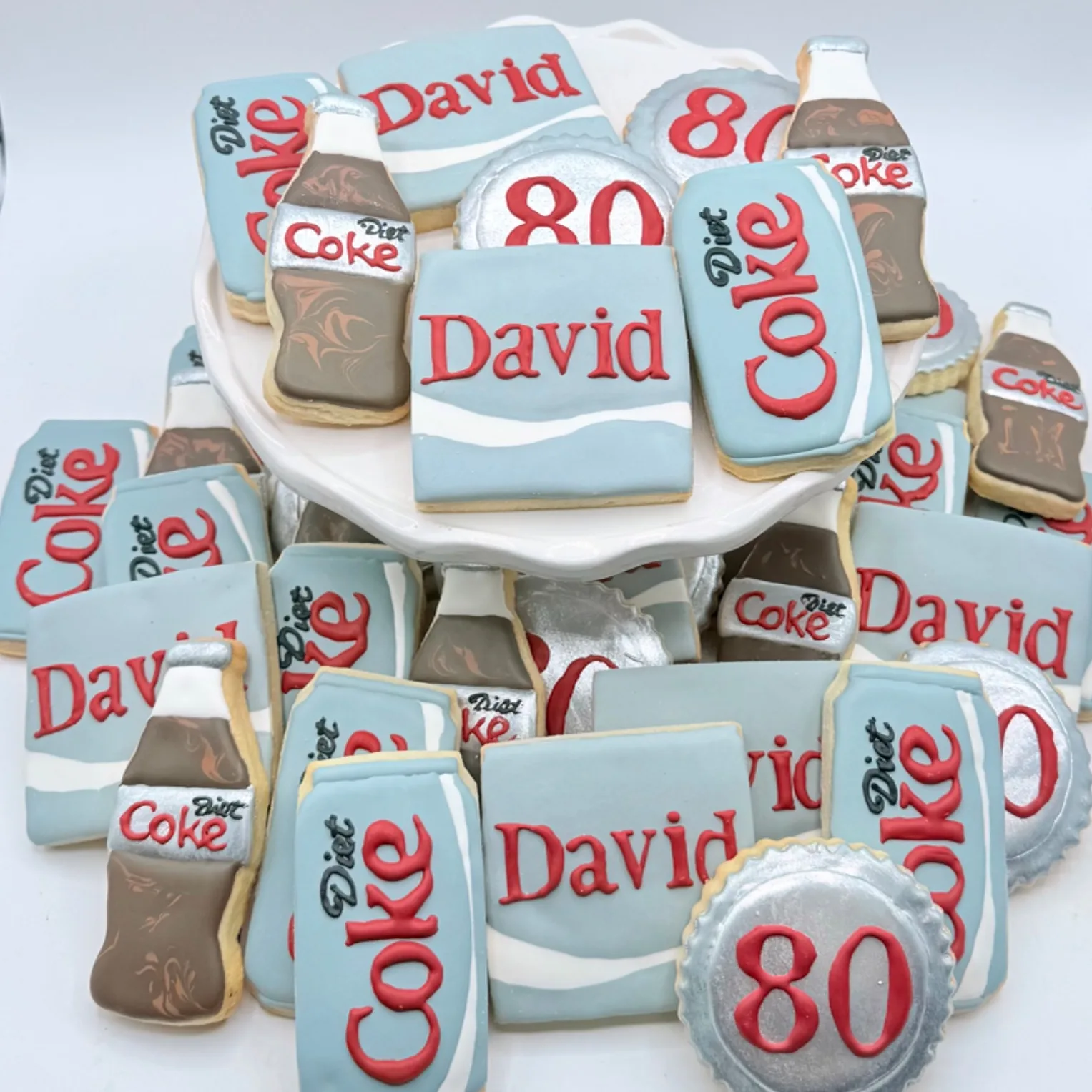 A tiered display of decorated cookies shaped like soda bottles, cans, and a silver bottle cap reading "80," alongside rectangular cookies piped with the name David, arranged on a white cake stand.