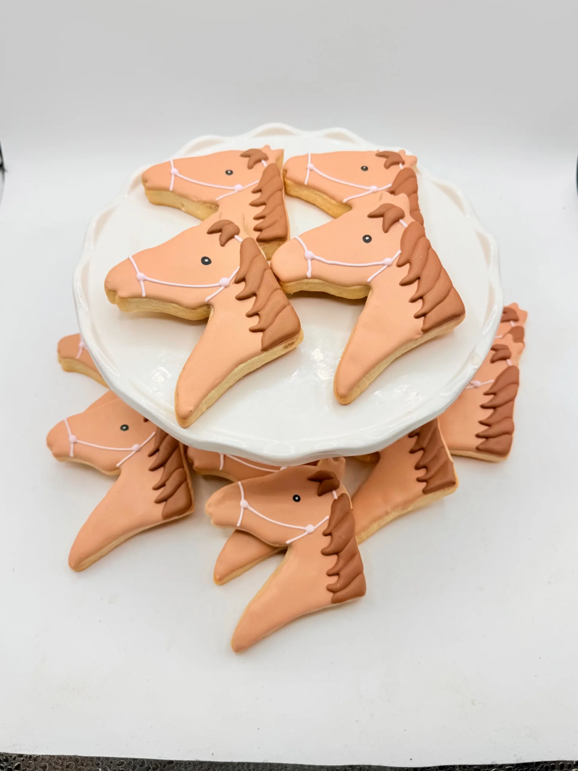 A cluster of horse-shaped sugar cookies with hand-piped brown manes, small painted eyes, and pink halter lines, arranged on a white scalloped plate and surrounding surface.