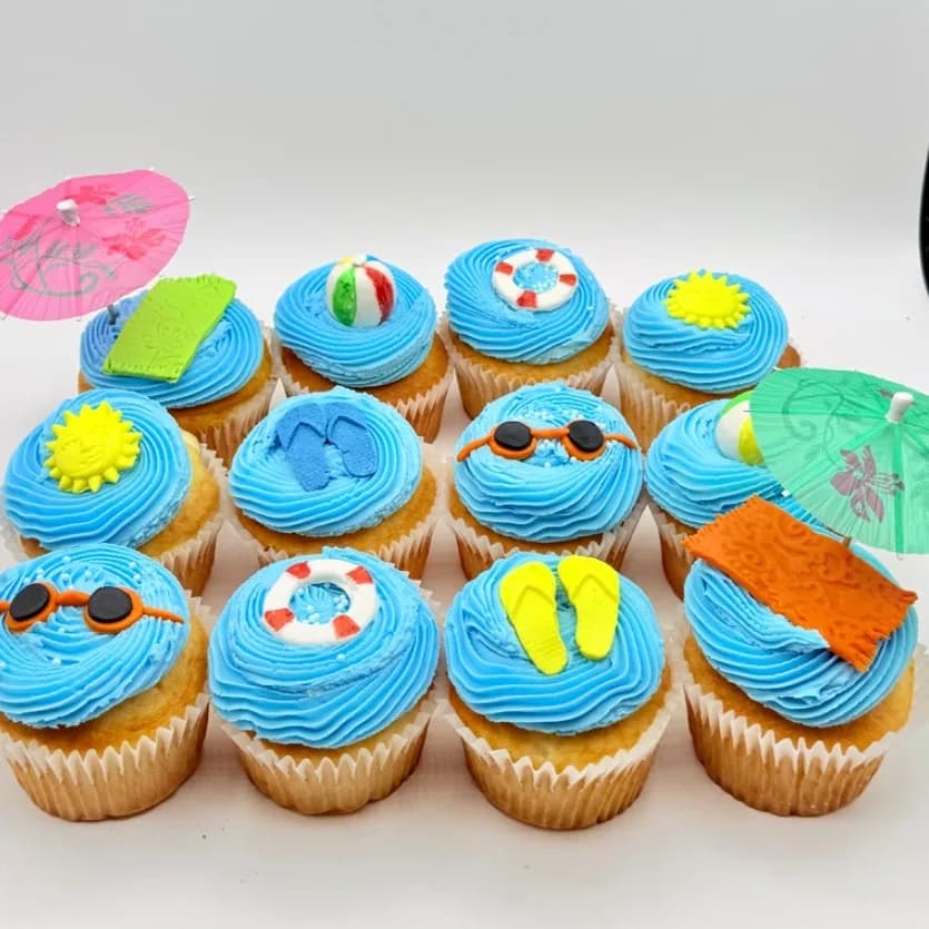 A dozen vanilla cupcakes with blue buttercream swirls topped with fondant sunglasses, flip flops, beach balls, pool floats, and towels, decorated with pink and green paper cocktail umbrellas.