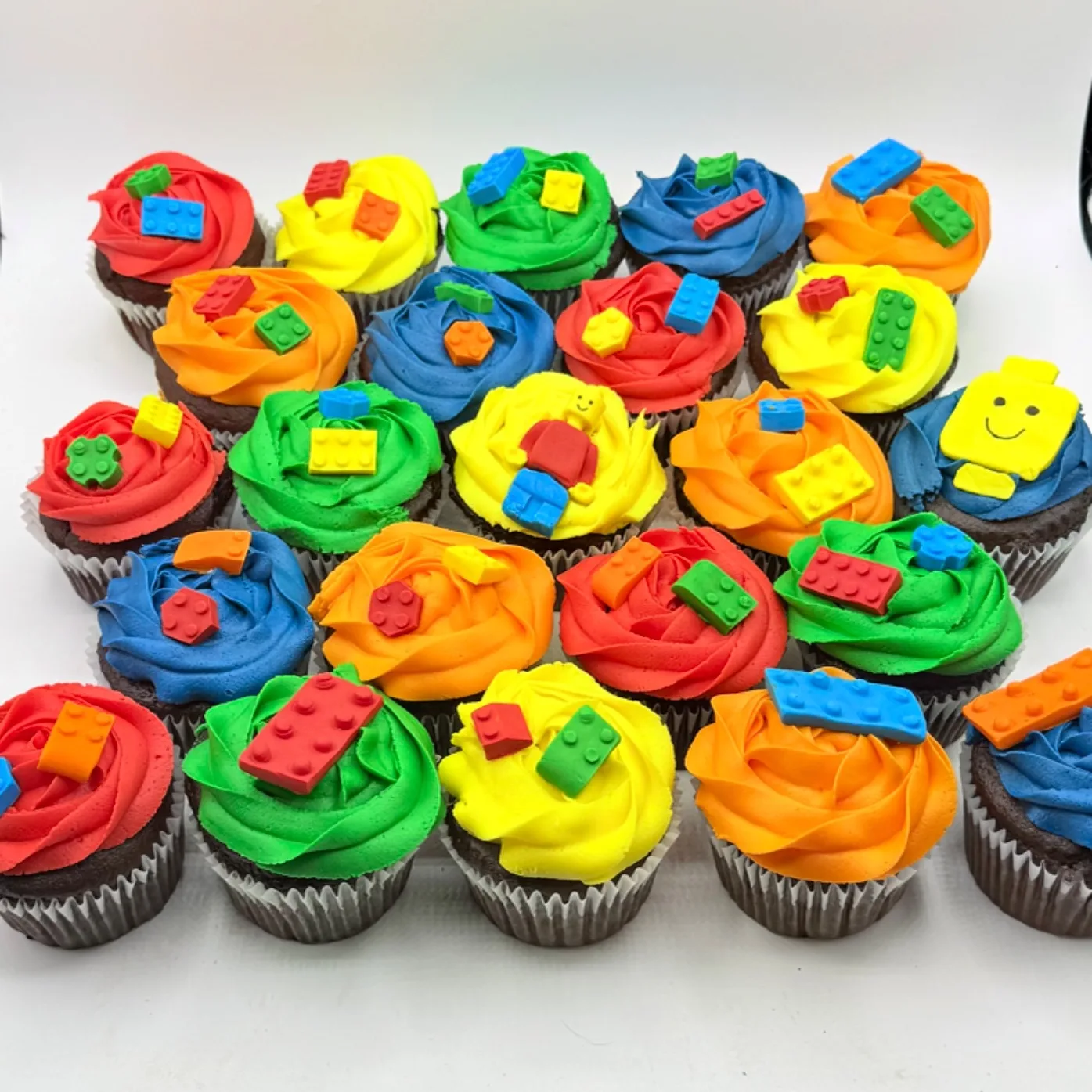 Twenty five chocolate cupcakes arranged in a grid, each topped with bright rosette swirls of red, yellow, green, blue or orange buttercream and decorated with colorful plastic-look brick toppers, including one full minifigure and one smiling block head topper.