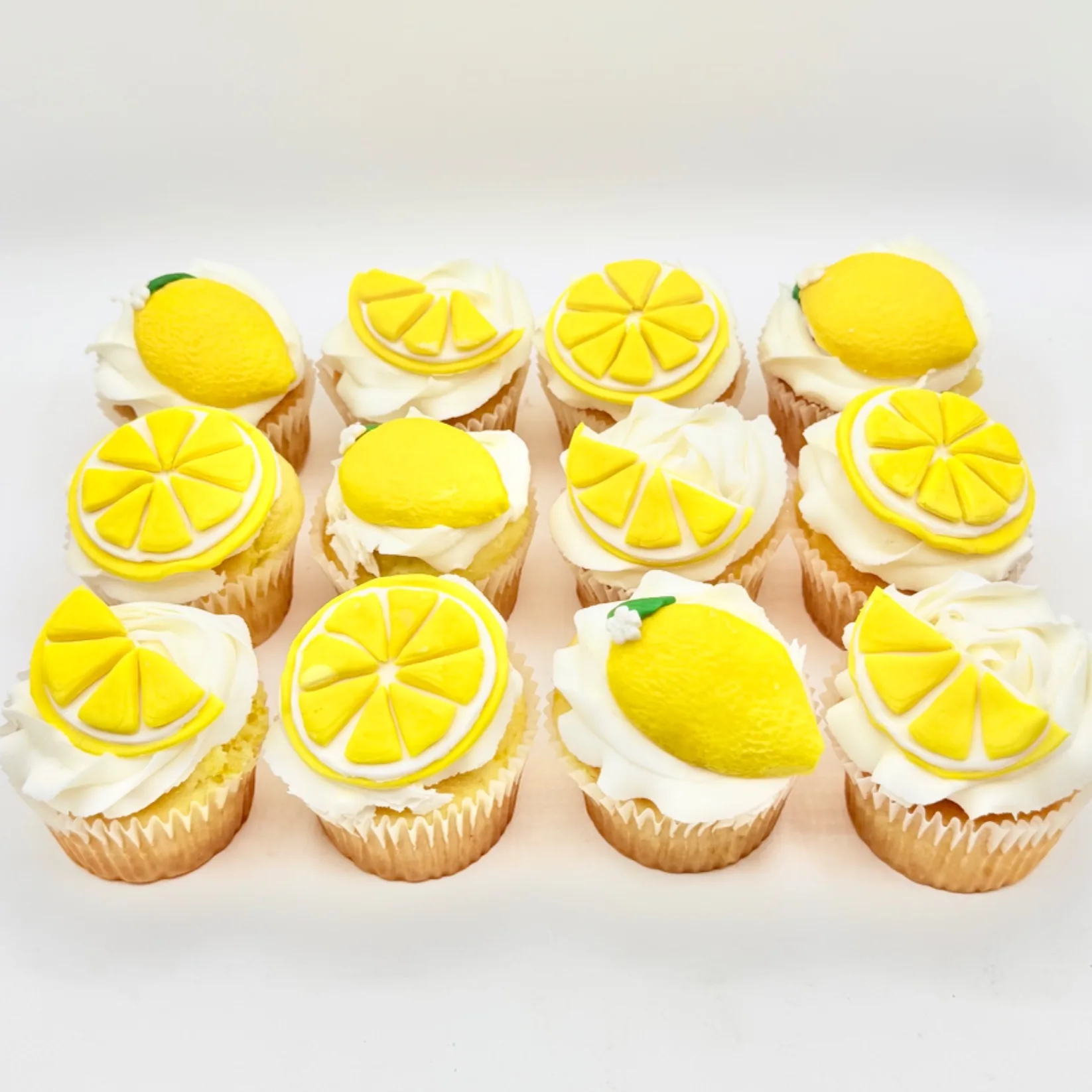 Twelve cupcakes with white buttercream swirls, each topped with a hand-piped fondant lemon slice or whole lemon in yellow with green leaf accents.