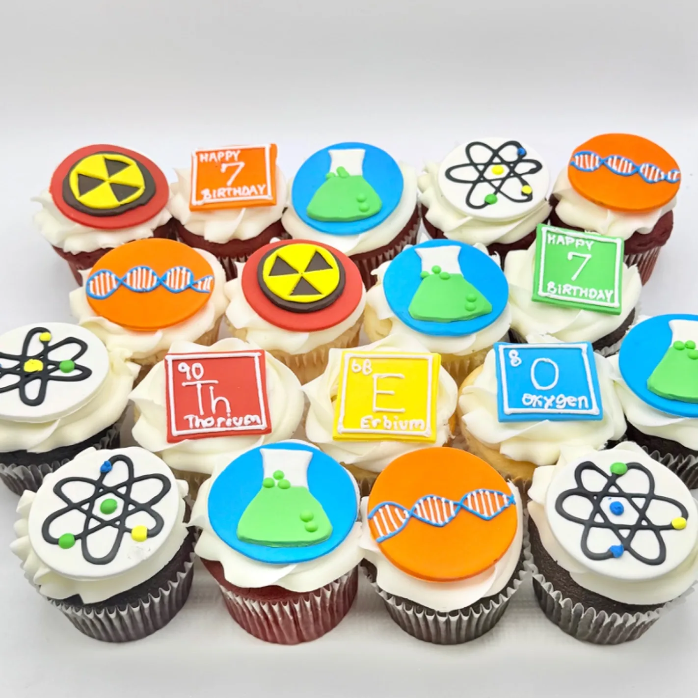 Rows of cupcakes topped with fondant discs and squares depicting atomic models, DNA strands, green beakers, radiation symbols, and periodic table elements spelling THEO and 7.