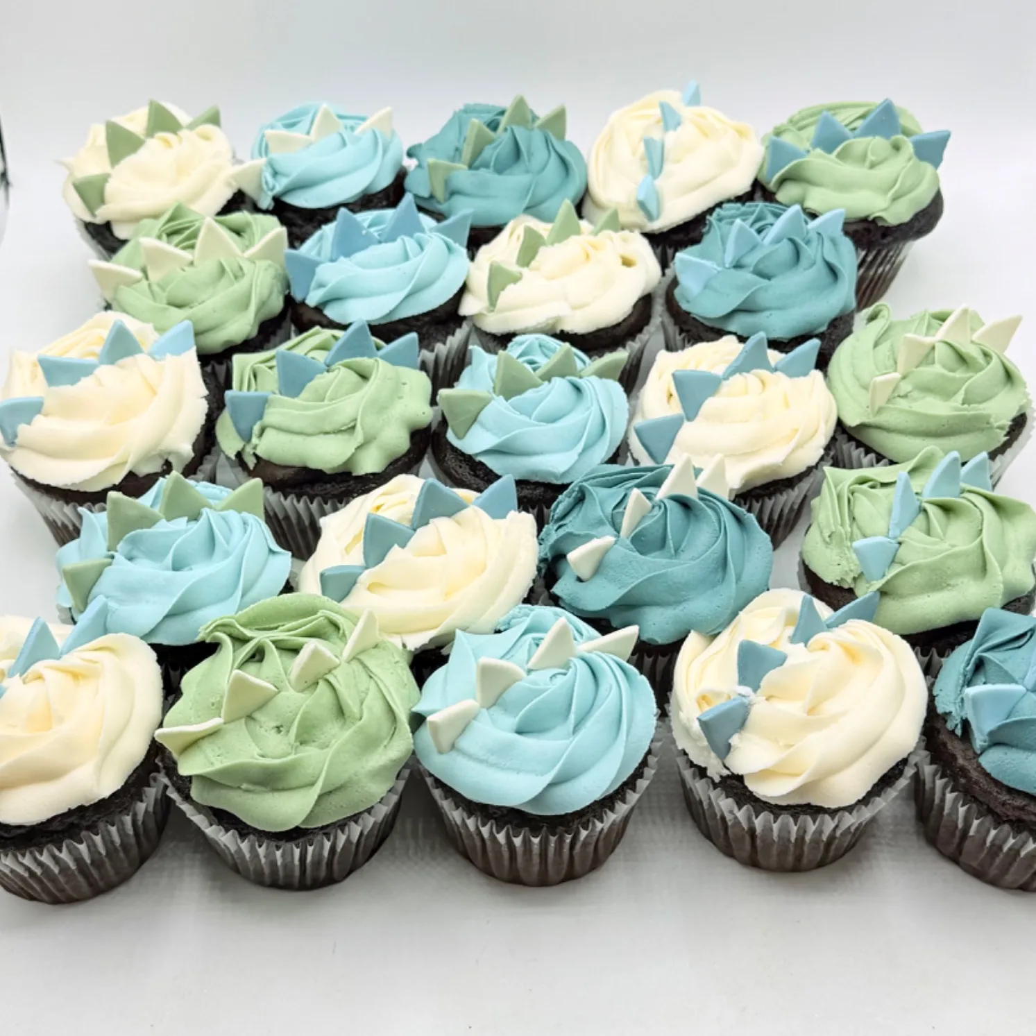 Rows of chocolate cupcakes topped with cream, sage green, and blue buttercream swirls, each finished with fondant triangle spikes resembling a dinosaur's back.