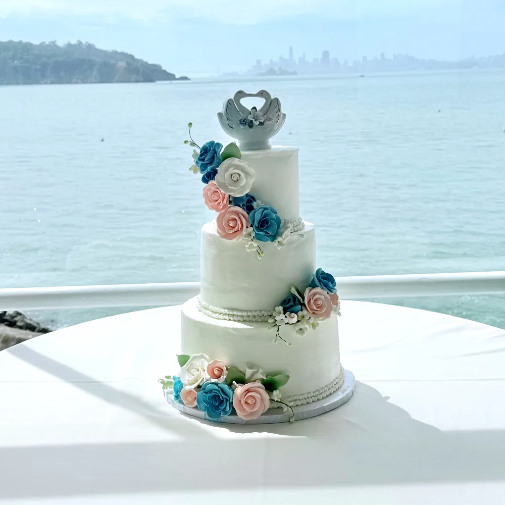 Katia three-tier white buttercream wedding cake with blue and peach sugar roses cascading down one side and a pale dove topper, set against San Francisco Bay, 2026