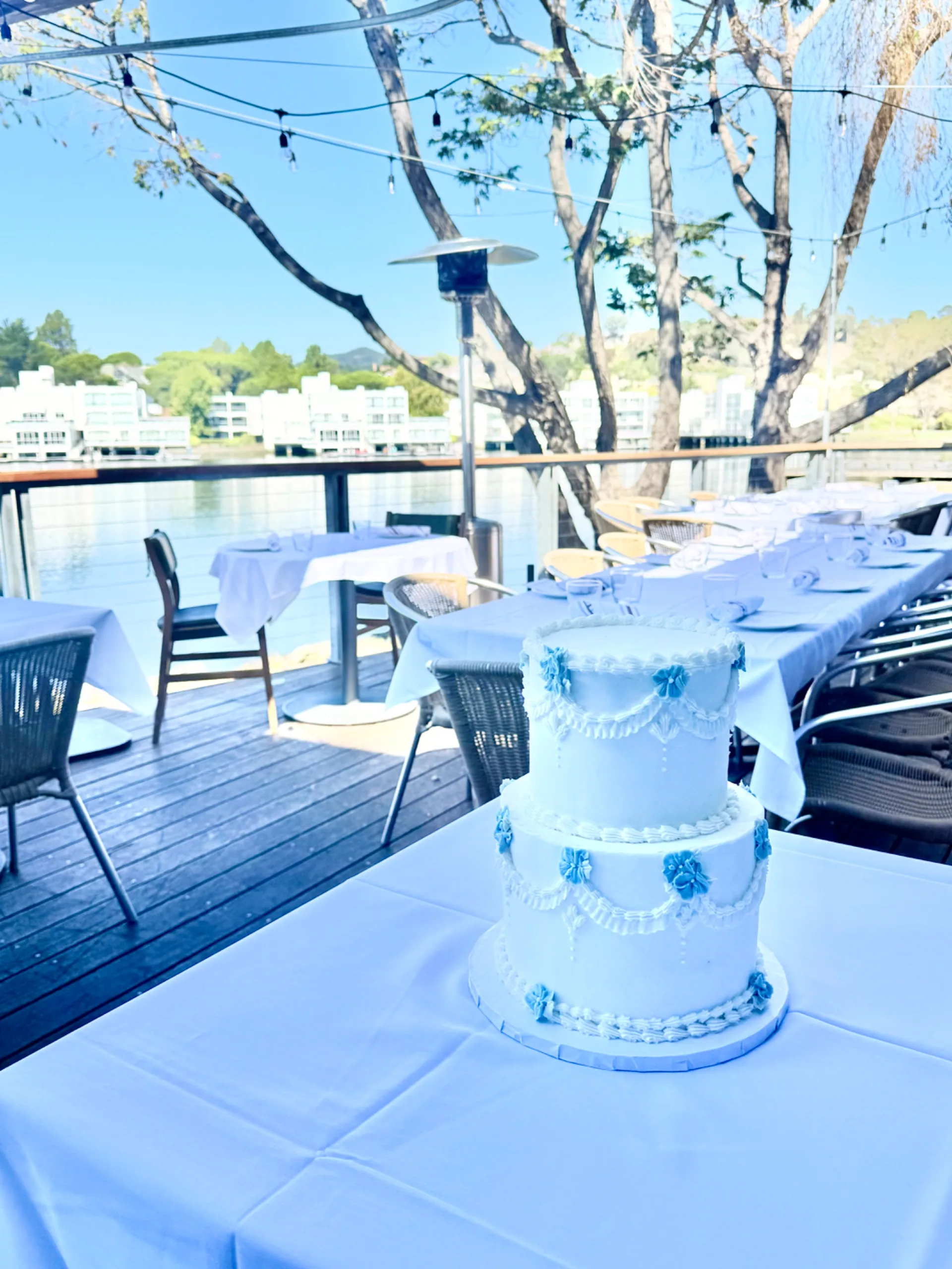 Annie two-tier white buttercream wedding cake with draped piping and dusty blue hand-piped florals, displayed on a lakeside restaurant deck with a long reception table set in the background, 2026