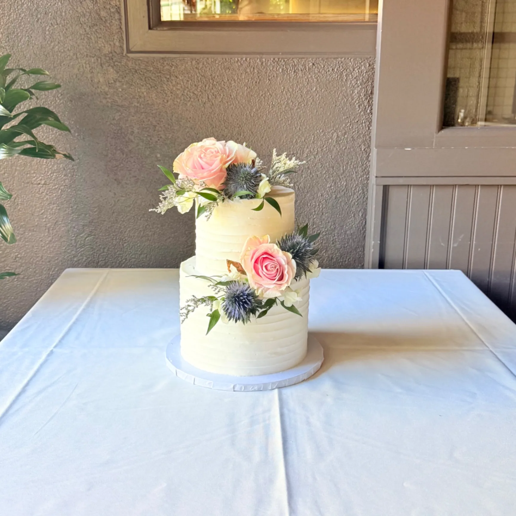 Elisa two tier round wedding cake with horizontally textured buttercream, topped and accented with blush pink roses, thistle, and greenery, displayed on a white linen covered table on an outdoor patio, 2026
