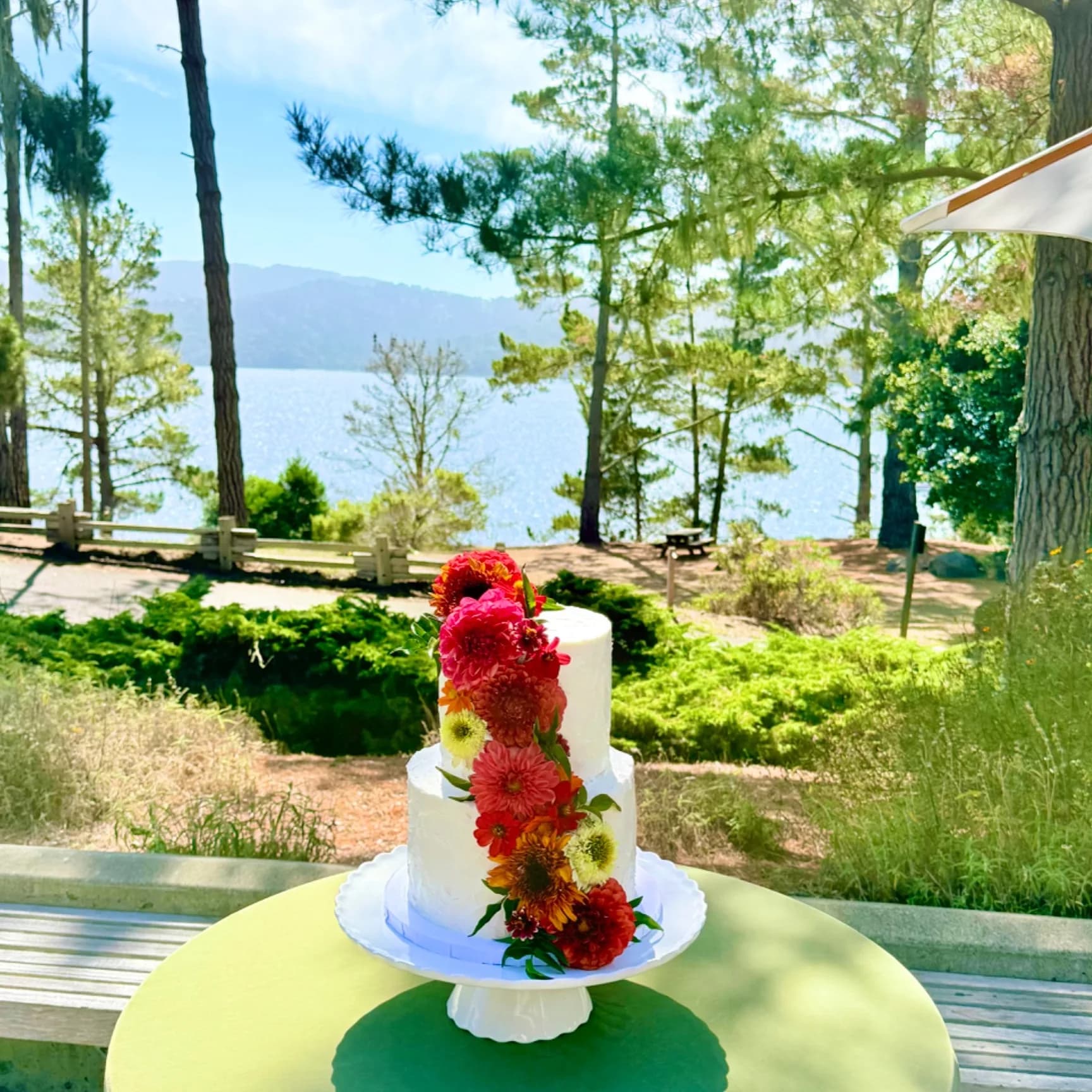 Rachel two-tier white textured buttercream wedding cake with a cascade of red, orange and yellow dahlias, zinnias and mums down one side, displayed on a cake stand on an outdoor table beside a lake framed by pine trees, 2026