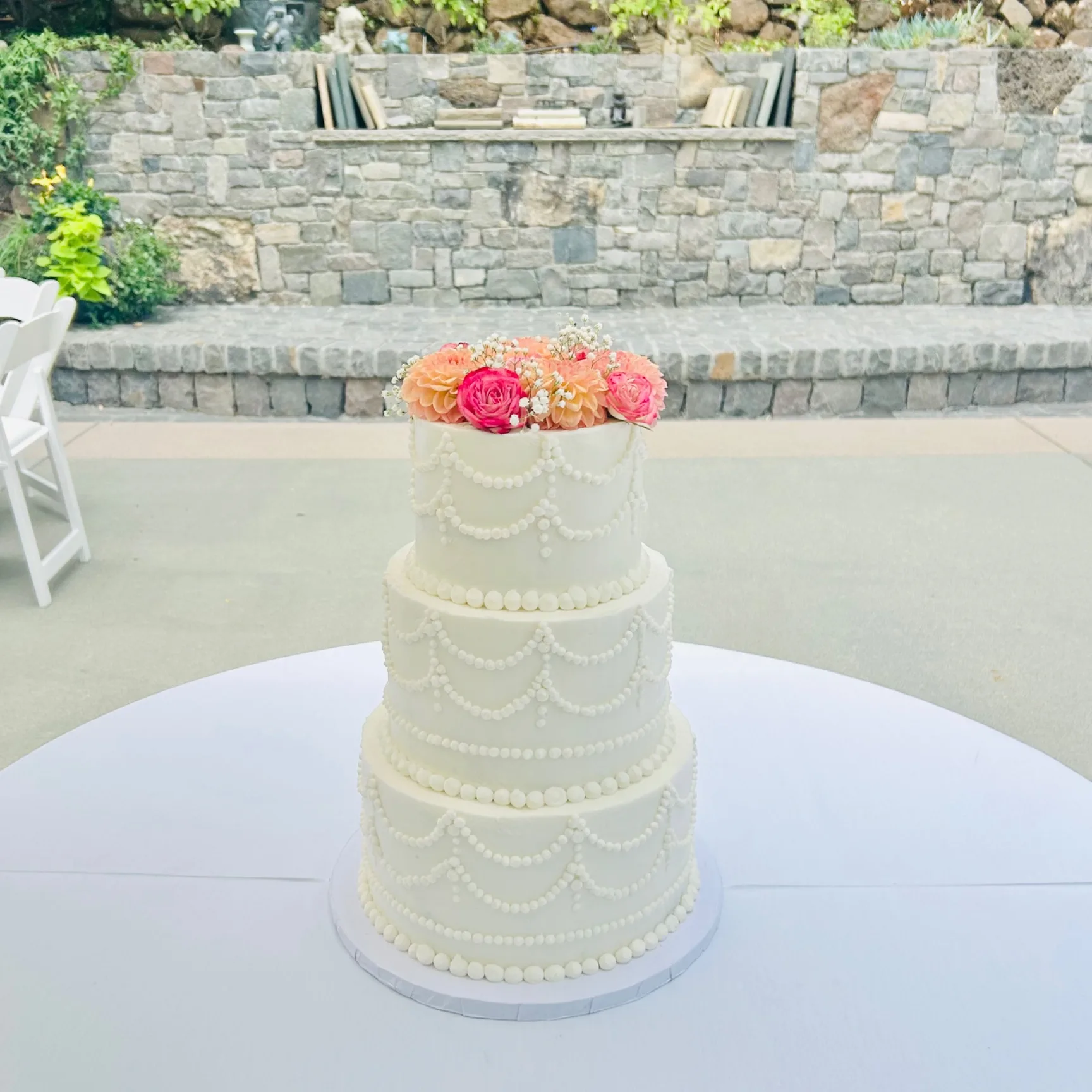 Carolyn three-tier white buttercream wedding cake with hand-piped pearl swag detailing on each tier, topped with fresh peach and pink garden roses, set outdoors against a stone wall, 2026